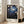 May include: Framed art print leaning against white wall next to beige sofa in bright room with large windows. Artwork features dark blue night sky with delicate white cherry blossoms on black branches, scattered white dots like stars or snow, and text reading "YAYOI KUSAMA" and Japanese characters with "TOKYO" below. Light wood frame, approximately 2-3 feet tall, positioned on light wooden floor.