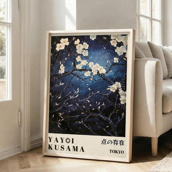 May include: Framed art print leaning against white wall next to beige sofa in bright room with large windows. Artwork features dark blue night sky with delicate white cherry blossoms on black branches, scattered white dots like stars or snow, and text reading "YAYOI KUSAMA" and Japanese characters with "TOKYO" below. Light wood frame, approximately 2-3 feet tall, positioned on light wooden floor.