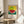 May include: Framed poster print featuring a colorful watermelon illustration with green rind and red flesh filled with black seeds, set against a yellow dotted background with blue and green horizontal bands below. Japanese characters and English text about courage appear beneath the artwork. The wooden-framed print sits on a round wooden side table next to a beige upholstered chair, with plant leaves visible in soft natural lighting.