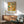 May include: Paul Klee abstract art poster with colorful geometric figures in yellow, orange, teal and red tones, displayed in thin wooden frame above mid-century modern walnut credenza. Scene shows sage green upholstered armchair with wooden arms, white ceramic table lamp, and sunlight casting shadows on white wall in contemporary living room setting.