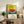 May include: Framed poster with colorful pointillism-style watermelon artwork against yellow dotted background, displayed on rustic wooden sideboard in cozy living room. Features Japanese characters and English text below the fruit illustration. Room setting includes white armchair, potted fern, ceramic vases, and books on weathered wood furniture.