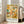 May include: Framed art poster leaning against white wall next to beige sofa in bright living room. Abstract artwork features hundreds of tiny colorful human figures in geometric shapes across warm yellow, orange, teal and green tones with vintage texture. White frame with black text below artwork showing artist name and dates. Natural sunlight streams through tall windows onto hardwood floors.