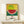 May include: Framed colorful watermelon artwork in pointillist style with yellow background, red fruit flesh with black seeds, and green rind. Japanese characters and English quote about courage below the image. Small white gift box with evergreen sprig sits on light wood surface beside the frame against white wall.