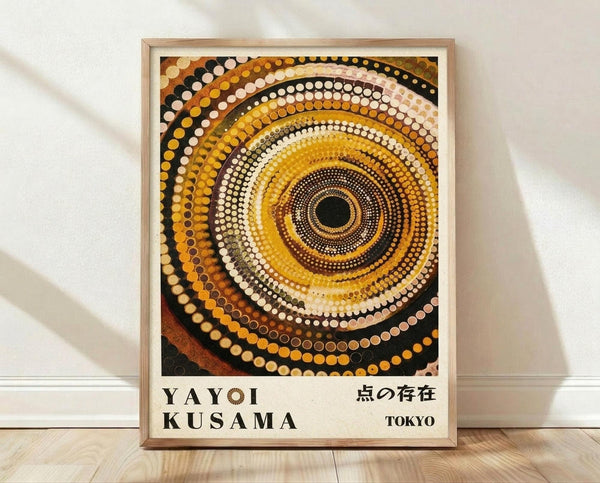May include: Framed art print featuring concentric circles of dots in warm earth tones - yellows, oranges, browns, and black - creating a hypnotic spiral pattern. Text reads "YAYOI KUSAMA" and Japanese characters with "TOKYO" below. Light wood frame sits on wooden floor against white textured wall with soft natural lighting casting shadows.
