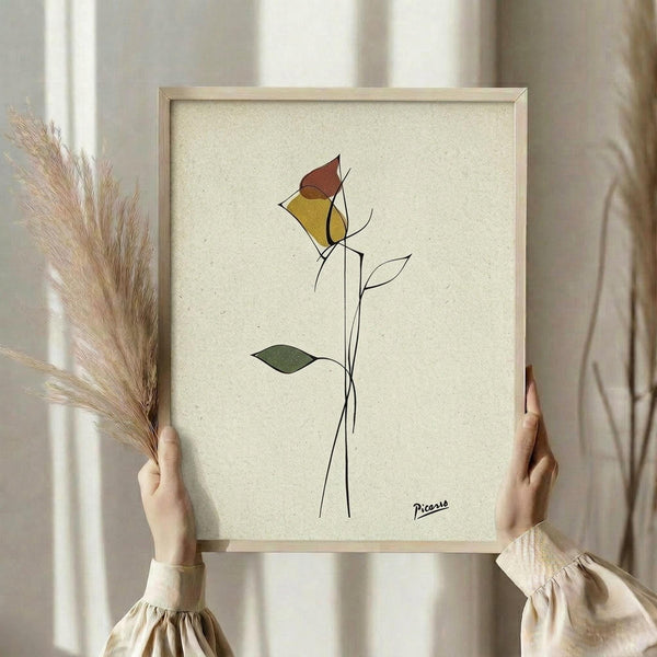 May include: Person in cream ruffled sleeves holding framed minimalist line art of a single rose with yellow and burgundy petals, green leaves on long stem, signed "Picasso" in bottom right corner. Cream textured background, light wood frame, dried pampas grass in left hand, soft natural lighting.