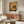 May include: Framed poster with concentric circles in warm earth tones - yellows, oranges, browns, and white dots radiating from black center, displayed above wooden sideboard in modern living room with cream chair, glass coffee table, and floor lamp