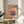 May include: Framed poster with colorful stack of pancakes in artistic style, featuring Japanese text and "itadakimasu" at bottom. Poster sits on light wood round side table next to beige upholstered chair. Plant leaves visible in corner, soft natural lighting creates shadows on white wall background.