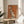 May include: Framed poster with orange background featuring an elegant sitting greyhound in cream and teal colors, displayed on a round wooden side table next to a beige upholstered chair. French text reads "L'élégance Ne Vieillit Pas" in script lettering. Natural sunlight streams across the minimalist room with white walls and a potted plant visible in the corner.