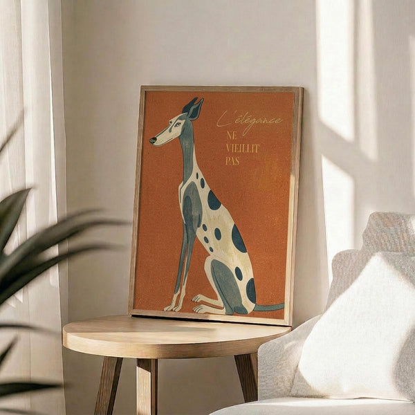 May include: Framed poster with orange background featuring an elegant sitting greyhound in cream and teal colors, displayed on a round wooden side table next to a beige upholstered chair. French text reads "L'élégance Ne Vieillit Pas" in script lettering. Natural sunlight streams across the minimalist room with white walls and a potted plant visible in the corner.