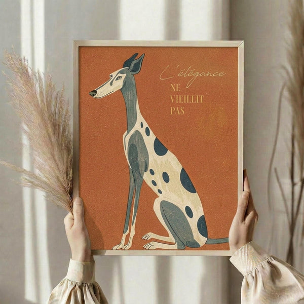 May include: Person in beige ruffled sleeves holding framed poster featuring stylized greyhound illustration on orange background. Dog has cream and teal spotted coat with long elegant neck and pointed ears. French text reads "L'elegance ne vieillit pas" in script and block letters. Natural wood frame, dried pampas grass visible on left side, neutral curtained background.