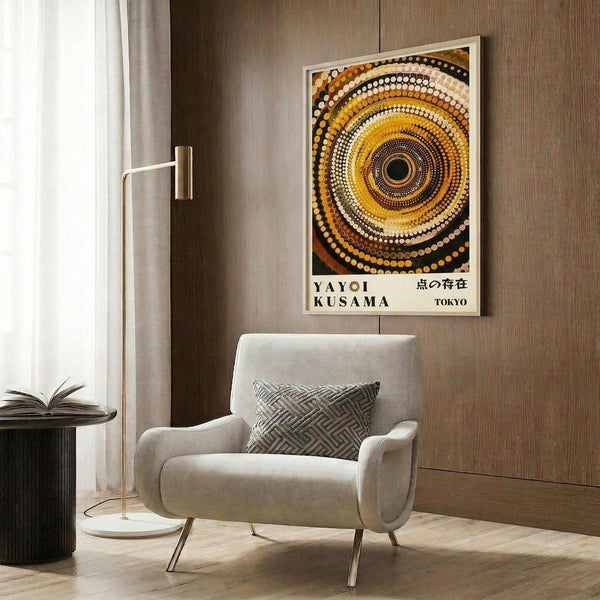 May include: A framed poster featuring concentric circles in warm tones of yellow, orange, brown, and white dots arranged in a hypnotic spiral pattern. The artwork displays "YAYOI KUSAMA" and Japanese text below. The print hangs on a wood-paneled wall above a cream upholstered armchair with geometric throw pillow, next to a brass floor lamp and dark round side table with open book.