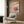 May include: Framed poster featuring colorful illustrated stack of pancakes with Japanese text and "itadakimasu" at bottom, displayed on wood-paneled wall above beige upholstered armchair with geometric patterned pillow. Modern brass floor lamp and dark round side table with open book complete the warm, contemporary living room setting with natural lighting from nearby window.