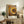 May include: Framed poster with concentric circles in warm tones of yellow, orange, brown, black and white dots creating a spiral pattern. Text reads "YAYOI KUSAMA" and Japanese characters with "TOKYO" below. Displayed on rustic wooden sideboard with books, ceramic vases, fern plant, and brown leather chair visible in cozy living room setting with cream walls.