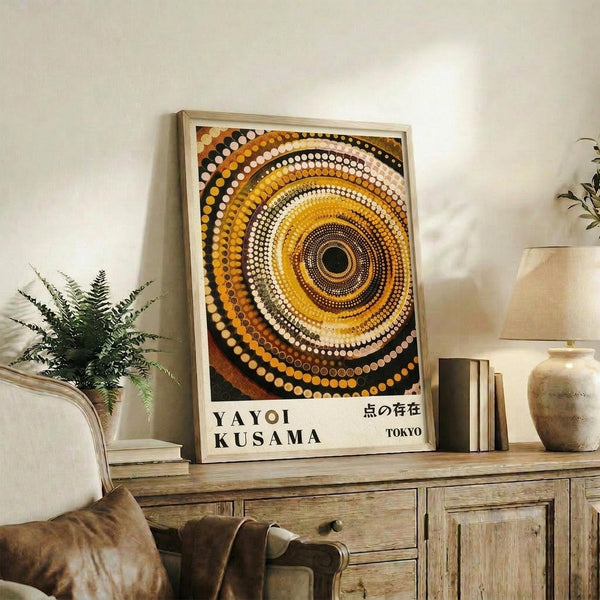 May include: Framed poster with concentric circles in warm tones of yellow, orange, brown, black and white dots creating a spiral pattern. Text reads "YAYOI KUSAMA" and Japanese characters with "TOKYO" below. Displayed on rustic wooden sideboard with books, ceramic vases, fern plant, and brown leather chair visible in cozy living room setting with cream walls.