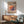 May include: Framed poster displaying colorful stack of pancakes with Japanese text on beige background, shown in mid-century modern living room setting with teal upholstered chair, wood sideboard, white ceramic lamp, and natural sunlight casting shadows on white wall.