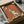 May include: Framed poster print featuring an elegant greyhound illustration in sage green and cream colors against a warm orange background, with French text reading "L'élégance Ne Vieillit Pas." The artwork is displayed in a black frame, nestled in brown cardboard packaging with protective tissue paper, sitting on a surface with neutral fabric and a dark glass object partially visible in the background.
