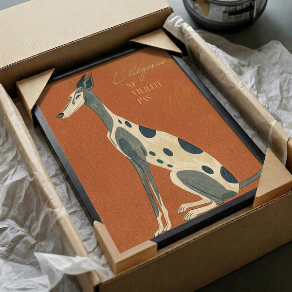 May include: Framed poster print featuring an elegant greyhound illustration in sage green and cream colors against a warm orange background, with French text reading "L'élégance Ne Vieillit Pas." The artwork is displayed in a black frame, nestled in brown cardboard packaging with protective tissue paper, sitting on a surface with neutral fabric and a dark glass object partially visible in the background.