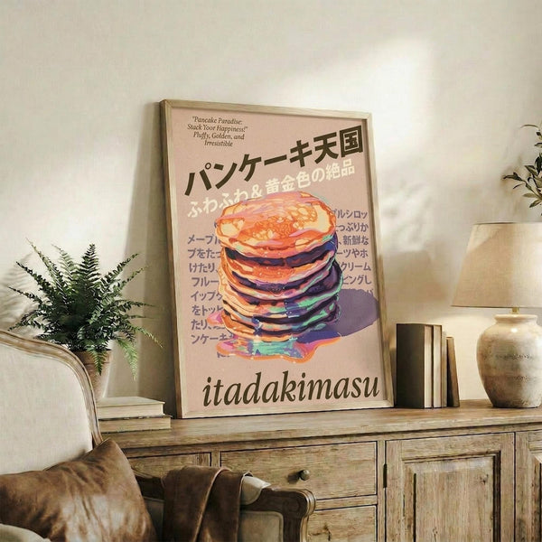 May include: Framed poster with colorful stack of pancakes artwork displayed on rustic wooden sideboard in cozy living room setting. Pink background with Japanese text and "itadakimasu" at bottom. Warm neutral decor includes potted fern, ceramic vase, leather chair, and books. Natural lighting creates soft shadows on cream walls.