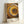 Framed poster featuring concentric circles of dots in warm earth tones - yellows, oranges, browns, and black - radiating from a central dark point. The dotted pattern creates a hypnotic spiral effect in the style of Japanese contemporary art. Text at bottom reads "YAYOI KUSAMA" on the left and Japanese characters with "TOKYO" on the right. The poster is displayed in a light wooden frame leaning against a white wall.