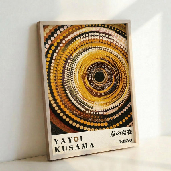 Framed poster featuring concentric circles of dots in warm earth tones - yellows, oranges, browns, and black - radiating from a central dark point. The dotted pattern creates a hypnotic spiral effect in the style of Japanese contemporary art. Text at bottom reads "YAYOI KUSAMA" on the left and Japanese characters with "TOKYO" on the right. The poster is displayed in a light wooden frame leaning against a white wall.