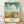 May include: Framed vintage-style travel poster featuring Savannah's historic district with tall Art Deco buildings, palm-lined streets, and puffy white clouds against turquoise sky. Poster shows "GEORGIA" in orange letters at top, displayed in light wood frame leaning against white wall on wooden floor with soft natural lighting.