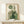 May include: Framed botanical abstract art print featuring green geometric shapes, circles, and plant motifs displayed on light wood surface next to small wrapped gift package with natural twine and sprig, against cream colored wall with soft natural lighting
