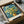 May include: Van Gogh print in black frame being unboxed from brown cardboard packaging with white tissue paper. The artwork shows yellow chrysanthemum flowers in a brown vase against swirling blue background. Print has white border with "VINCENT VAN GOGH" and "VAN GOGH MUSEUM, AMSTERDAM" text at bottom. Wooden dowels visible as protective packaging elements.