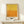May include: Abstract art poster with yellow to orange gradient color field in light wood frame, displayed on white wooden shelf beside small wrapped gift with green sprig, against cream wall with natural lighting