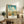 May include: Vintage-style travel poster featuring Georgia with Savannah Historic District cityscape in warm blues, yellows and oranges, displayed in light wood frame on rustic wooden sideboard alongside fern plant, pottery vase, and books in cozy living room setting with beige chair and neutral walls