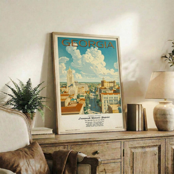May include: Vintage-style travel poster featuring Georgia with Savannah Historic District cityscape in warm blues, yellows and oranges, displayed in light wood frame on rustic wooden sideboard alongside fern plant, pottery vase, and books in cozy living room setting with beige chair and neutral walls