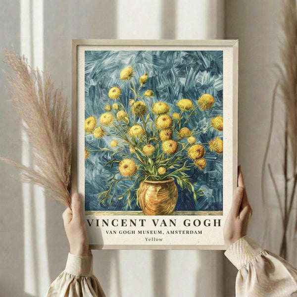 May include: Woman in cream ruffled sleeves holding framed Van Gogh museum poster print featuring yellow chrysanthemum flowers in golden vase against blue brushstroke background. Dried pampas grass visible beside frame. Neutral indoor setting with soft natural lighting and beige curtains.