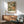 May include: Framed botanical art print featuring large detailed flowers in green, cream, and orange tones hanging above a mid-century modern wooden credenza. Gray upholstered armchair with wooden frame sits nearby, white ceramic table lamp on credenza. Warm sunlight creates diagonal shadows on white wall. Print shows intricate floral design with dotted patterns and flowing organic shapes in vintage Japanese art style.