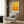 May include: Yellow to orange gradient abstract art print in white frame hanging on wood paneled wall above beige upholstered armchair with geometric patterned pillow. Modern brass floor lamp and dark round side table with open book visible. Warm neutral toned room setting with natural lighting from window with curtains.