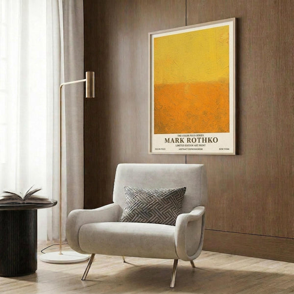 May include: Yellow to orange gradient abstract art print in white frame hanging on wood paneled wall above beige upholstered armchair with geometric patterned pillow. Modern brass floor lamp and dark round side table with open book visible. Warm neutral toned room setting with natural lighting from window with curtains.