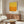 May include: Yellow and orange abstract color field art print displayed above wooden sideboard in modern living room. White upholstered chair and glass coffee table visible. Print shows textured gradient from bright yellow at top transitioning to deep orange below, housed in thin light wood frame against neutral wall.