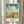 May include: Framed vintage travel poster featuring aerial view of Savannah Historic District with white colonial buildings, red clay roofs, palm-lined streets under dramatic blue sky with puffy white clouds. Poster displays "GEORGIA" in orange letters at top with descriptive text below. Light wood frame hangs on white wall with soft natural lighting and curtains visible in background.