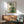 May include: Abstract botanical wall art in wooden frame above mid-century modern credenza. Green and cream artwork features geometric circles, plant stems, and linear patterns. Sage green upholstered chair with wooden arms sits beside credenza with white ceramic lamp. Warm sunlight creates dramatic shadows across white wall and wooden furniture surfaces.