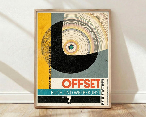 May include: Framed vintage Bauhaus poster with geometric design featuring concentric circles, curved shapes in yellow, black, sage green, and coral colors. German text reads "OFFSET BUCH UND WERBEKUNST HEFT 7 1926". Light wood frame displayed against white wall on wooden floor with soft natural lighting and shadows.