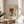May include: Framed vintage-style art print propped on round wooden side table next to beige upholstered chair. Artwork features minimalist illustration of hands holding wine glass with red wine, stylized eyes peering from below, rendered in muted earth tones on cream background. Text reads "Plaisir des yeux" at bottom. Natural light creates soft shadows across white wall and plant leaves visible in corner.