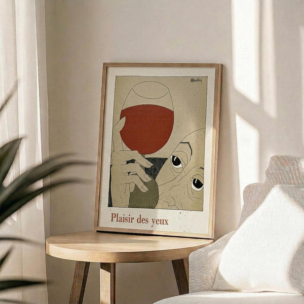 May include: Framed vintage-style art print propped on round wooden side table next to beige upholstered chair. Artwork features minimalist illustration of hands holding wine glass with red wine, stylized eyes peering from below, rendered in muted earth tones on cream background. Text reads "Plaisir des yeux" at bottom. Natural light creates soft shadows across white wall and plant leaves visible in corner.