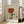 May include: Framed vintage-style poster with beige textured background showing stylized hands holding wine glass with red wine, large eyes gazing upward, and French text "Plaisir des yeux" below. Light wood frame leaning against white wall next to beige upholstered chair in bright room with windows.