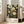 May include: Framed botanical art print leaning against white wall next to beige sofa in bright living room. Black background features golden yellow flowers, circular seed heads, feathery grasses, and white dotted patterns. White bottom section displays artist name and exhibition details. Natural wood frame, approximately 2-3 feet tall.