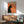 May include: Vintage-style poster featuring black cat silhouette with yellow eye smoking against orange background, displayed in wooden frame above mid-century modern credenza. Scene shows teal upholstered armchair with wooden arms, white ceramic table lamp, and round coffee table in bright, naturally-lit room with white walls.
