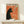 May include: Vintage-style poster with orange background featuring black silhouette of a cat with pointed ears and green eye, smoking a cigarette with wispy smoke. French text reads "Le Chat Nocturne" at top. Displayed in thin wooden frame on light wood shelf next to small wrapped gift box against white wall.