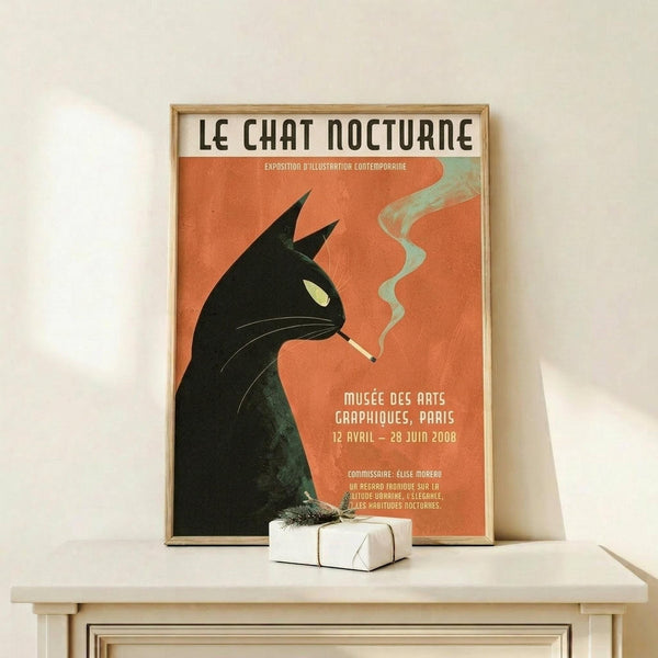 May include: Vintage-style poster with orange background featuring black silhouette of a cat with pointed ears and green eye, smoking a cigarette with wispy smoke. French text reads "Le Chat Nocturne" at top. Displayed in thin wooden frame on light wood shelf next to small wrapped gift box against white wall.