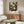 May include: Framed botanical art poster with dark background featuring cream and yellow circular flowers, dandelions, and foliage displayed above a mid-century modern walnut credenza in a minimalist living room with curved white chair, glass coffee table, and natural lighting from nearby window.