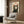 May include: Framed poster of a black and white tuxedo cat with yellow eyes and open mouth saying "Meow" in red text, displayed on wood paneled wall above a beige upholstered armchair with geometric patterned pillow. Modern living room setting with brass floor lamp, side table with open book, and white curtains visible.