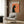 May include: Framed vintage-style poster featuring black cat silhouette smoking against orange background with French text, displayed above cream upholstered chair with geometric pillow in modern living room with wood paneling, floor lamp, and side table with open book.