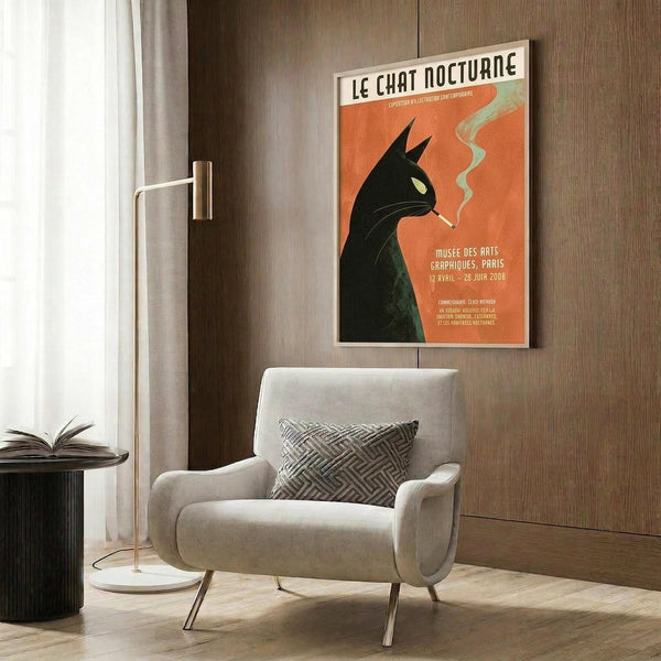 May include: Framed vintage-style poster featuring black cat silhouette smoking against orange background with French text, displayed above cream upholstered chair with geometric pillow in modern living room with wood paneling, floor lamp, and side table with open book.