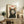 May include: Framed vintage-style poster featuring a black and white tuxedo cat with bright yellow eyes and orange ear interiors, with red "Meow" text above, displayed on rustic wooden sideboard alongside books, ceramic vase, and fern plant in cozy living room setting with beige armchair and neutral walls